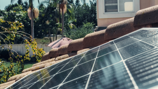 "Close-up of rooftop solar panels on a tiled roof with a residential building and greenery in the background.