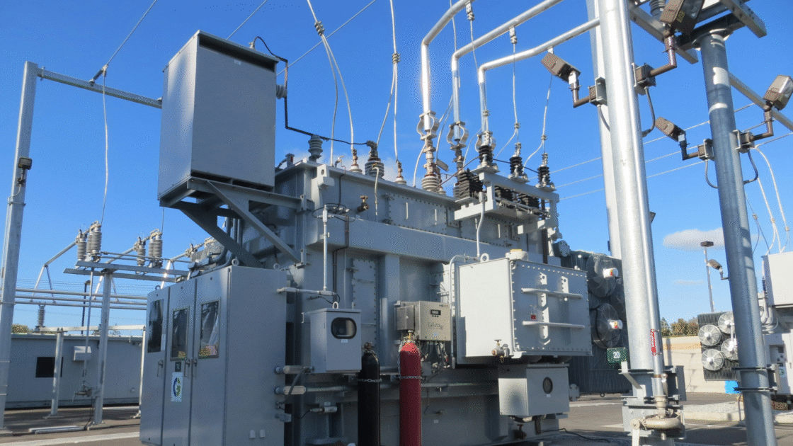 A large industrial power transformer in a substation, featuring gray metal housing, insulators, bushings, cooling fans, and overhead power lines against a clear blue sky. Red and black gas cylinders are visible at the base.