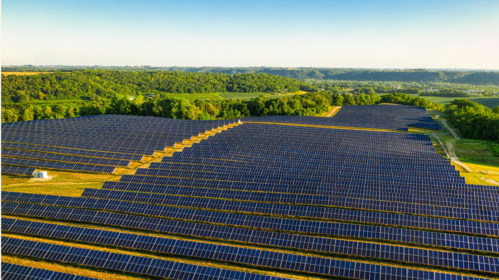 Aerial view of a large solar farm surrounded by greenery under a clear blue sky.