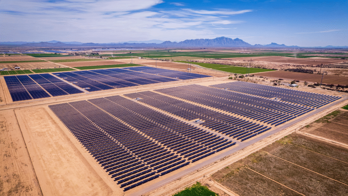 Aerial view of a large solar farm with rows of photovoltaic panels installed on flat, dry land, surrounded by agricultural fields and distant mountain ranges under a clear sky.