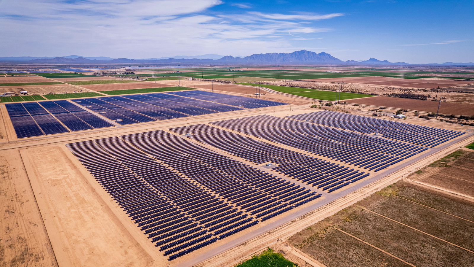 Aerial view of a large solar farm with rows of photovoltaic panels installed on flat, dry land, surrounded by agricultural fields and distant mountain ranges under a clear sky.