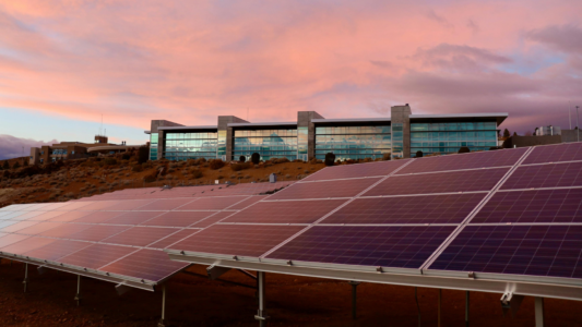 Ground mounted solar panels in the foreground with a modern glass front building on a hillside