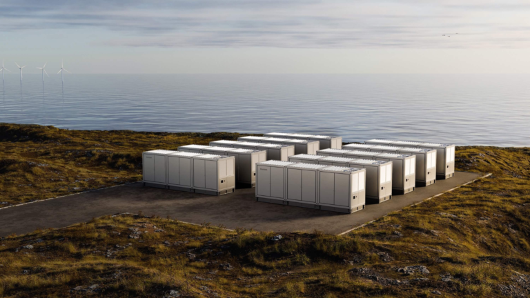 A cluster of rectangular battery storage units arranged on a cleared platform overlooking calm coastal waters.