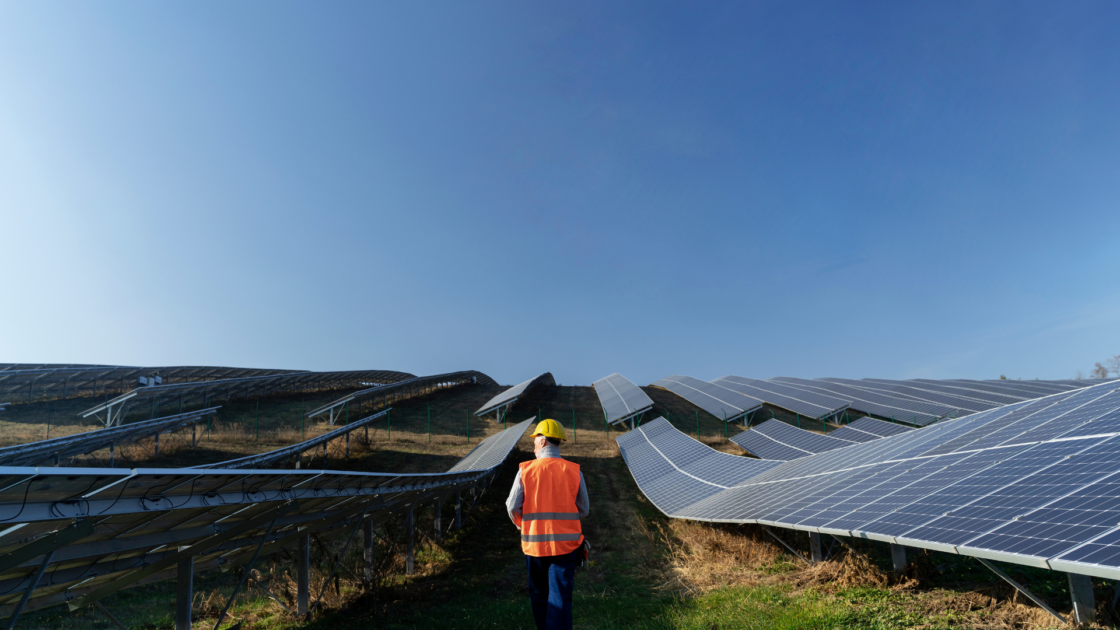 Worker wearing a safety helmet and high visibility vest walks between rows of ground mounted solar panels at a utility scale solar power plant under a clear sky.