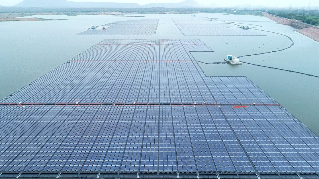 Aerial view of a floating solar installation with dense rows of photovoltaic panels spread across a calm reservoir, with surrounding hills, embankments, and maintenance platforms visible in the distance.