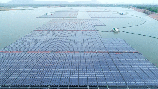 Aerial view of a floating solar installation with dense rows of photovoltaic panels spread across a calm reservoir, with surrounding hills, embankments, and maintenance platforms visible in the distance.