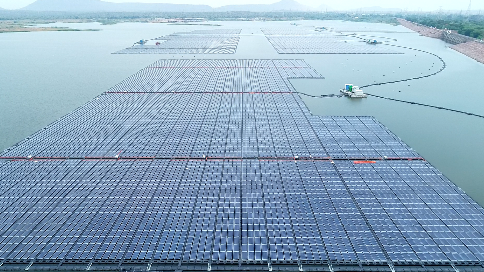 Aerial view of a floating solar installation with dense rows of photovoltaic panels spread across a calm reservoir, with surrounding hills, embankments, and maintenance platforms visible in the distance.