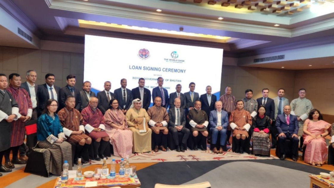 Officials and delegates from the Royal Government of Bhutan and the World Bank pose for a group photograph at the loan signing ceremony for the Dorjilung Hydroelectric Power Project. A banner reading "Loan Signing Ceremony between Royal Government of Bhutan and the World Bank" is visible on the screen behind them.