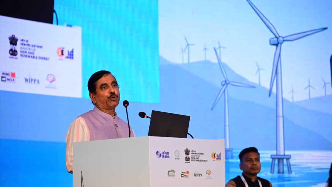 Union Minister for New and Renewable Energy Pralhad Joshi addresses delegates at the Global Wind Day Conference 2026 in Goa, with wind turbines displayed on the event backdrop during the launch of the WT-MARUT wind supply chain portal.